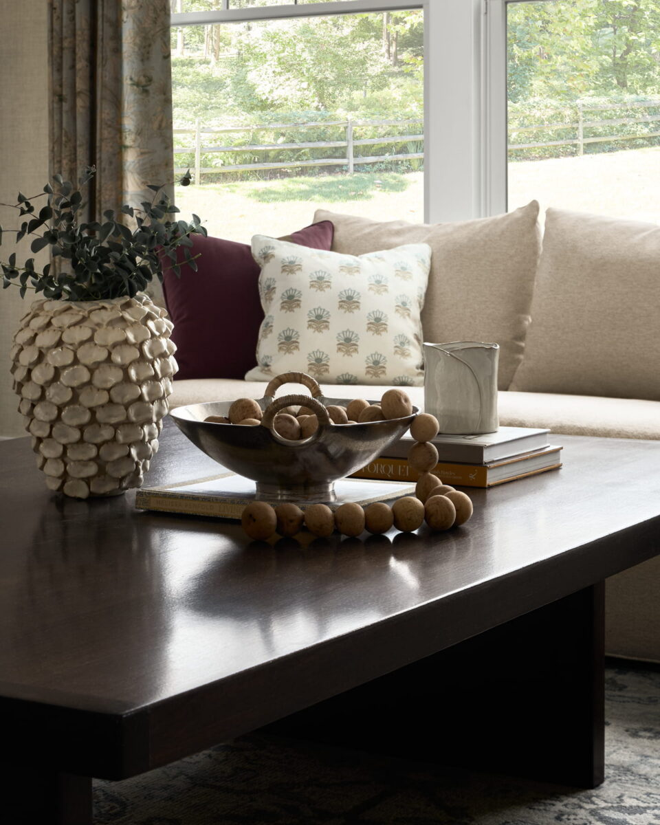 A dark wood coffee table holds decorative beads, a bowl, books, a textured vase with greenery, and a candle. In the background, a beige couch with pillows sits in front of a bright window with a view of trees.