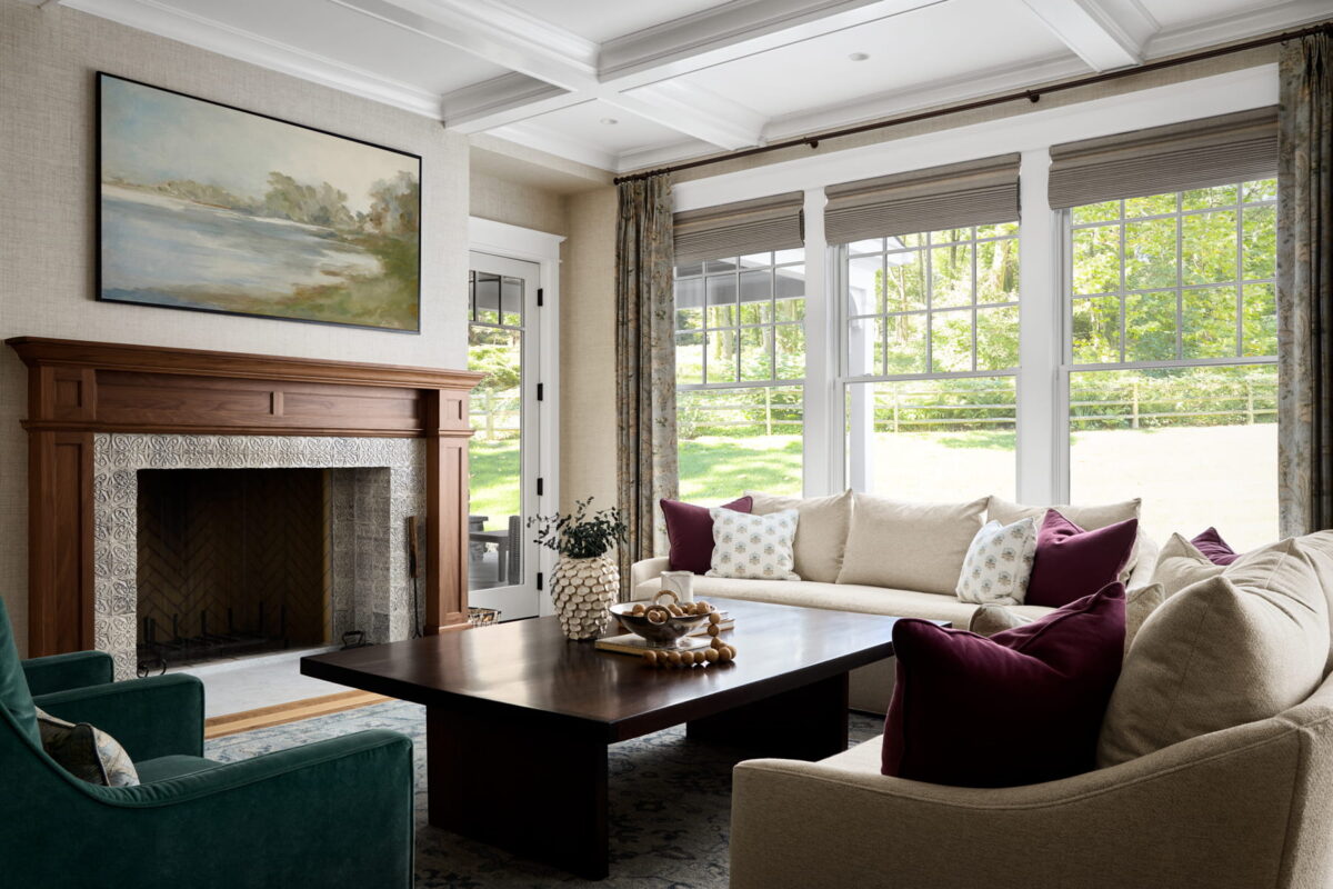 Bright living room with large windows, beige sofa, green armchair, dark wood coffee table, fireplace with wood mantel, landscape painting above, and decorative pillows. Sunlight streams in, highlighting cozy, elegant décor.