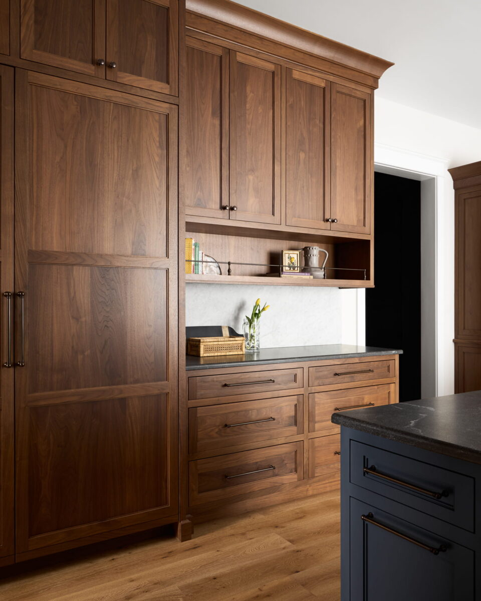 A modern kitchen with tall wooden cabinets, drawers, and open shelves, a marble backsplash, a vase with flowers, and part of a dark-colored island. The floor is light wood.