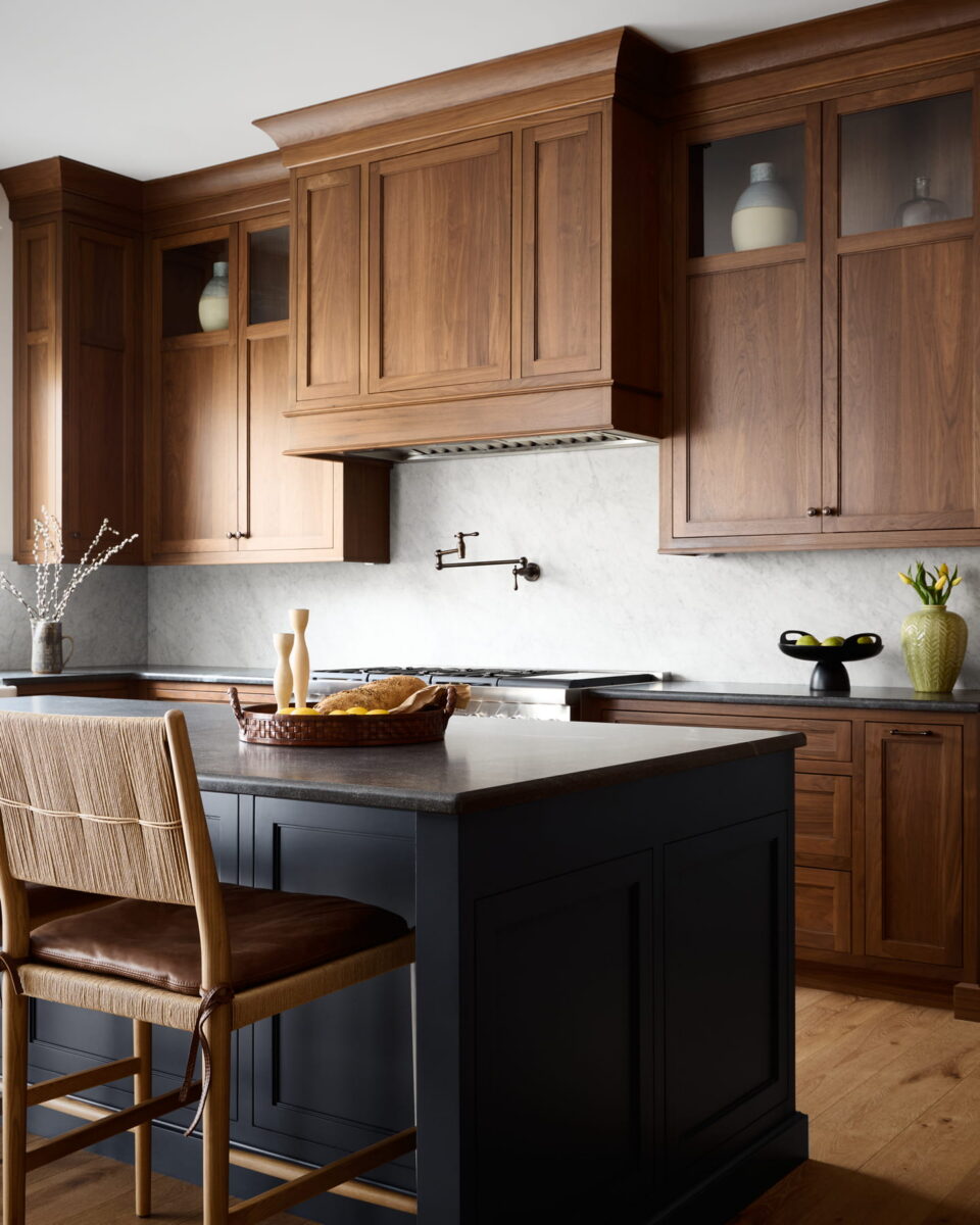 Modern kitchen with wooden cabinets, marble backsplash, black island, wicker chair, and simple decor including vases and a fruit bowl. Warm lighting and a clean, inviting atmosphere.