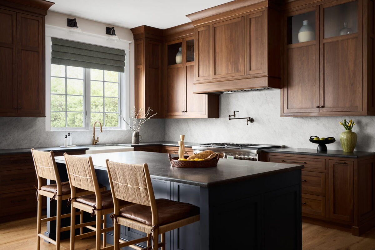 A modern kitchen with dark wood cabinets, marble backsplash, a large island with three chairs, a window with a roman shade, and decorative items on the counters.
