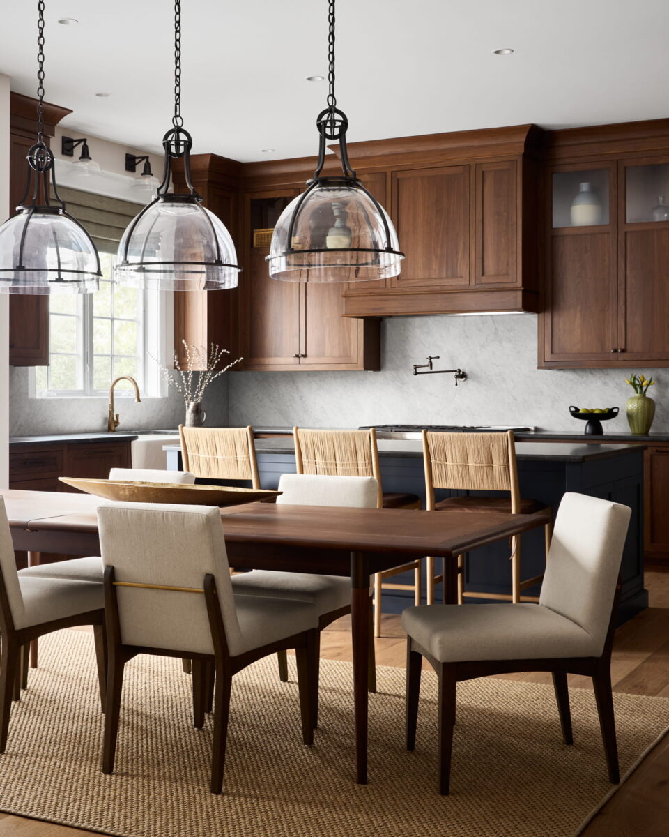 A modern kitchen with wooden cabinets, a marble backsplash, a large island, glass pendant lights, and a dining table with six chairs on a woven rug. Natural light streams through the window.