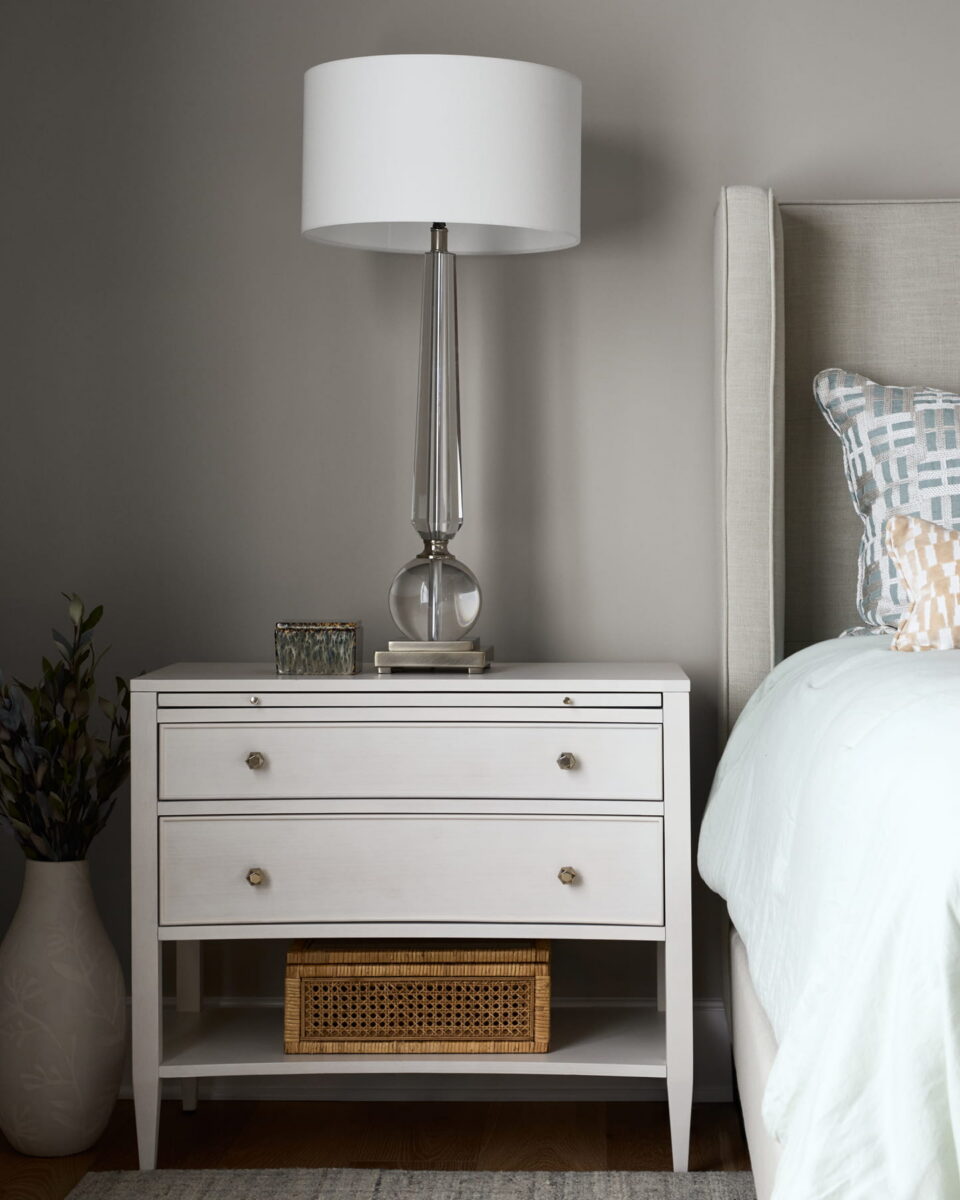 A modern bedroom features a white nightstand with two drawers, a glass lamp, a small plant, and books, next to a bed with a light blue comforter and decorative pillows.