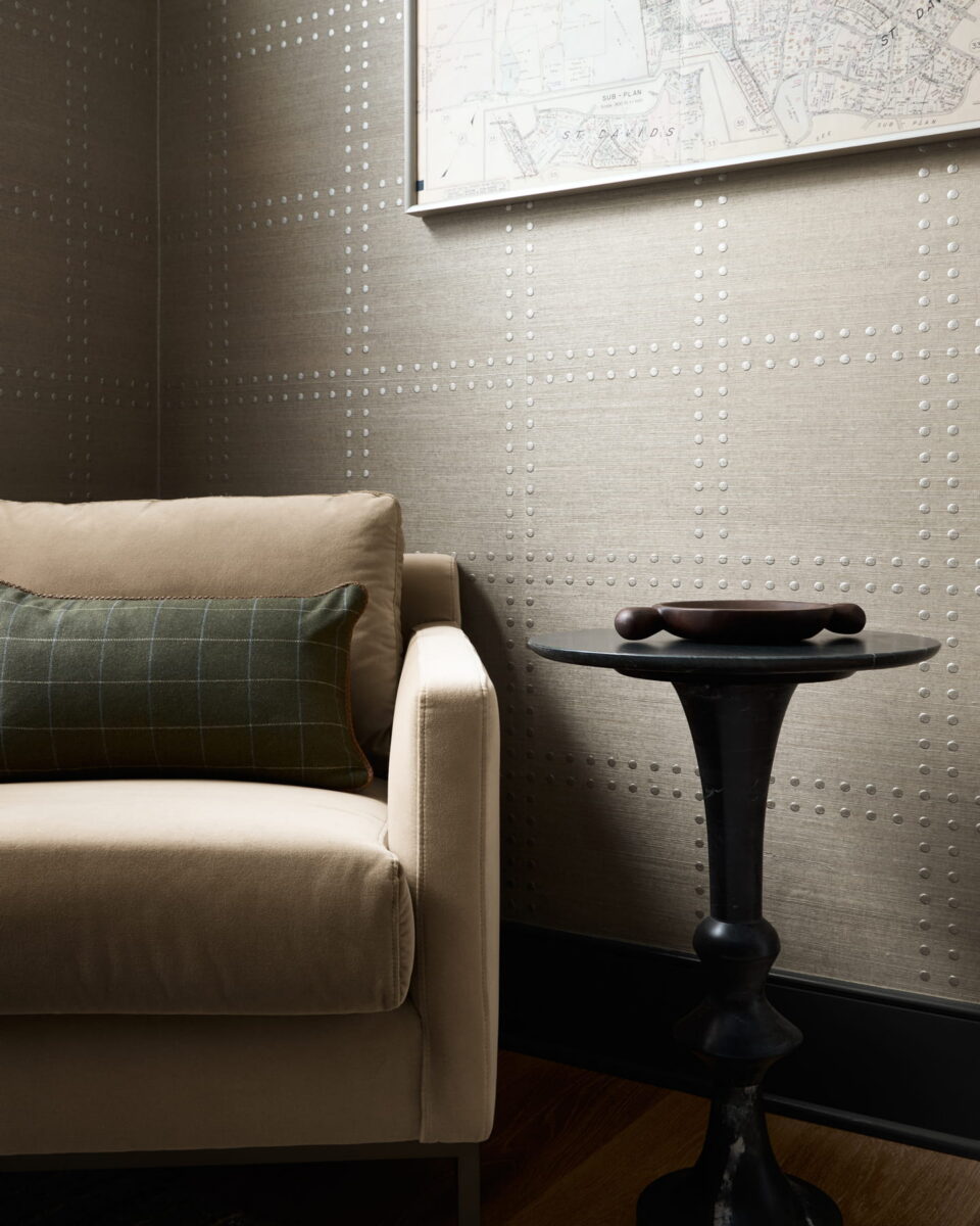 Beige armchair with a green plaid pillow next to a black round side table holding a decorative bowl. Behind them, textured wallpaper and a framed map are partially visible on the wall.