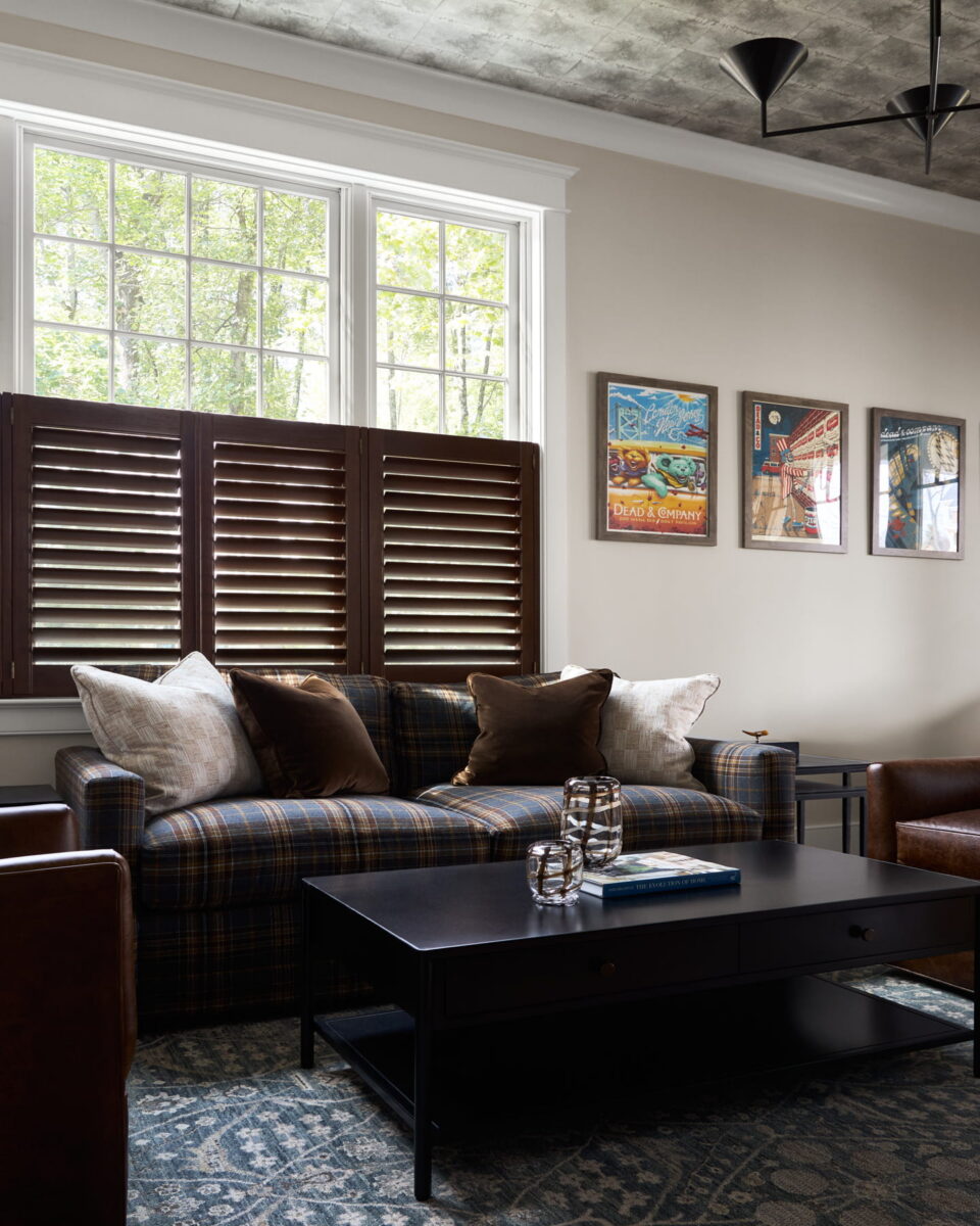 A living room with a plaid sofa, brown and cream pillows, wooden shutters, framed wall art, a dark coffee table with books, and patterned rug. Large windows let in natural light.