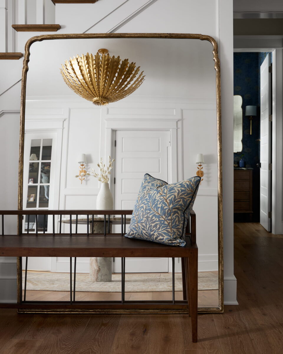 A large gold-framed mirror reflects a white wall with doors, a gold chandelier, and a wooden bench with a blue patterned pillow. The floor is wood, and a vase with branches sits on a small table.