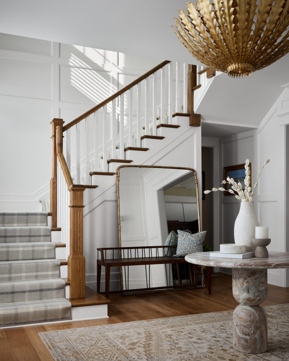 A bright entryway with a staircase, plaid carpet runner, large gold mirror, wooden bench with pillows, white vase with branches, and a round table under a gold chandelier.