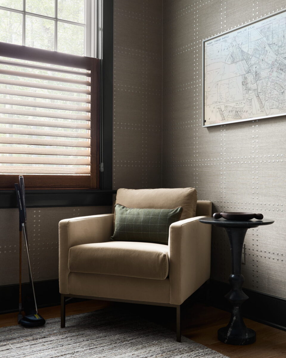 A beige armchair with a green cushion sits by a window with wooden blinds. A small black side table holds a bowl, and two golf clubs lean against the wall. A framed map hangs above the chair.