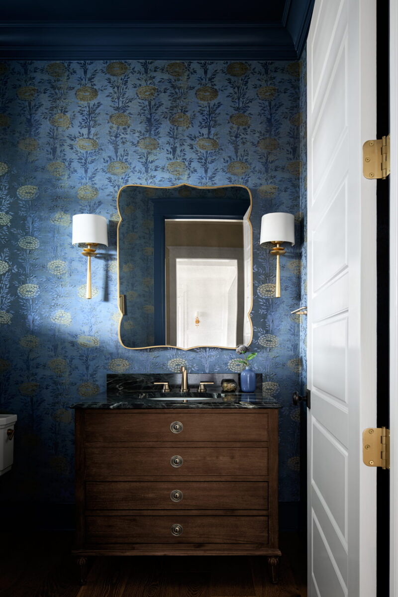A bathroom with blue floral wallpaper, a wooden vanity with a marble top, a gold-framed mirror, two wall sconces, and a white door slightly open, reflecting hallway light.