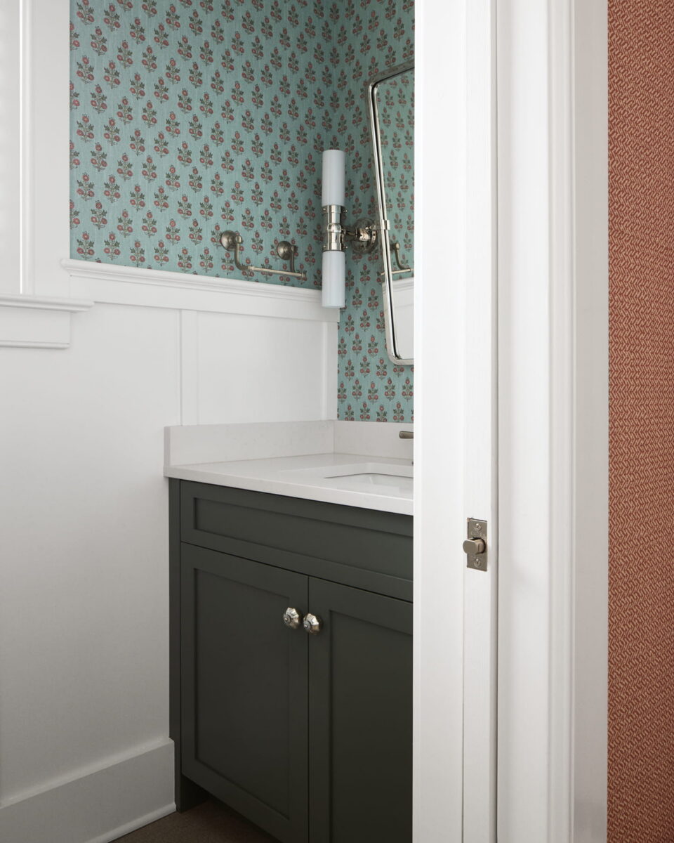 A small bathroom with green cabinets, white countertop, floral-patterned blue wallpaper, a rectangular mirror, and a wall-mounted light fixture next to the sink. The door is partially open.