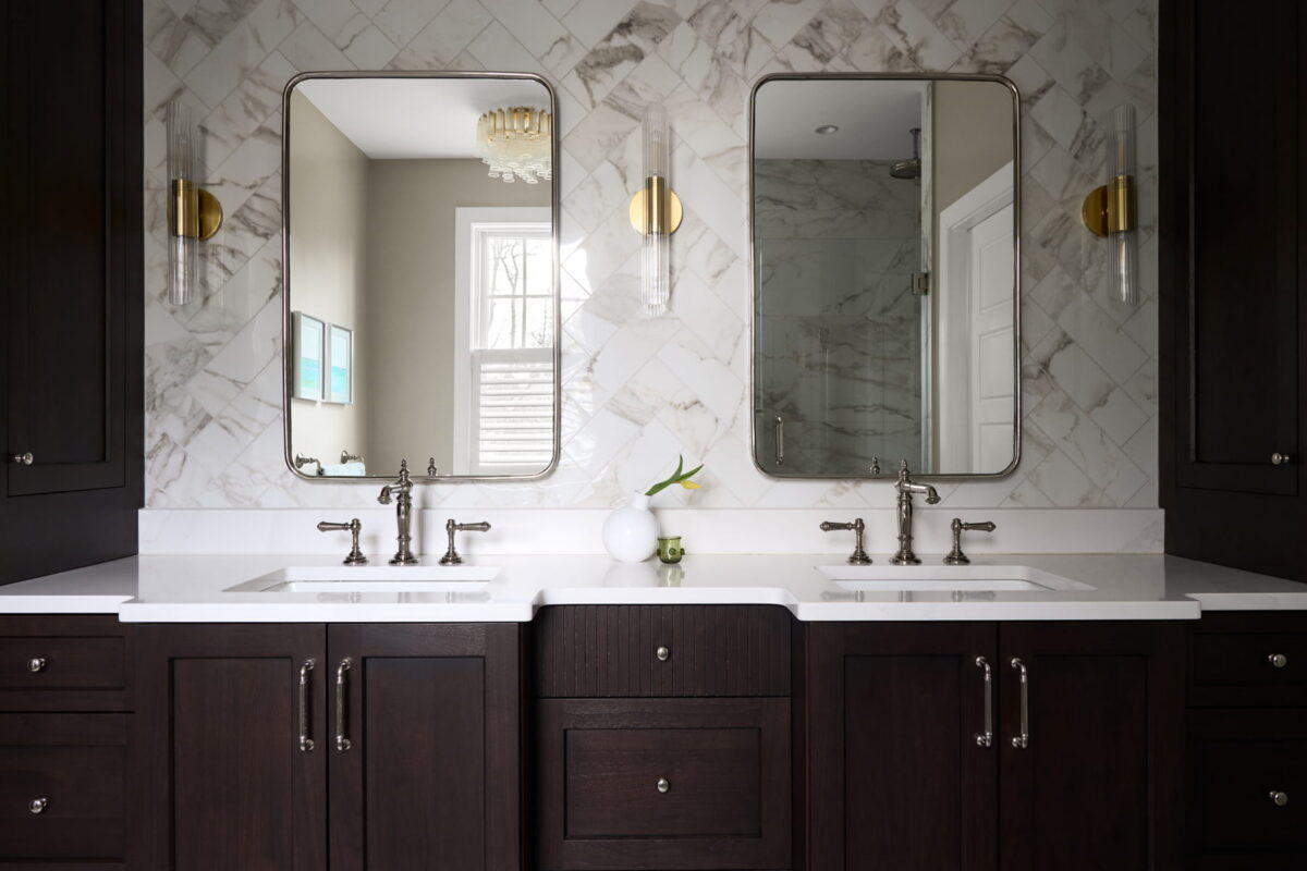 Modern bathroom with two sinks, dark wood cabinets, white countertops, two mirrors, wall sconces, and a small vase with a white flower on the counter. Marble tile wall and window reflect natural light.
