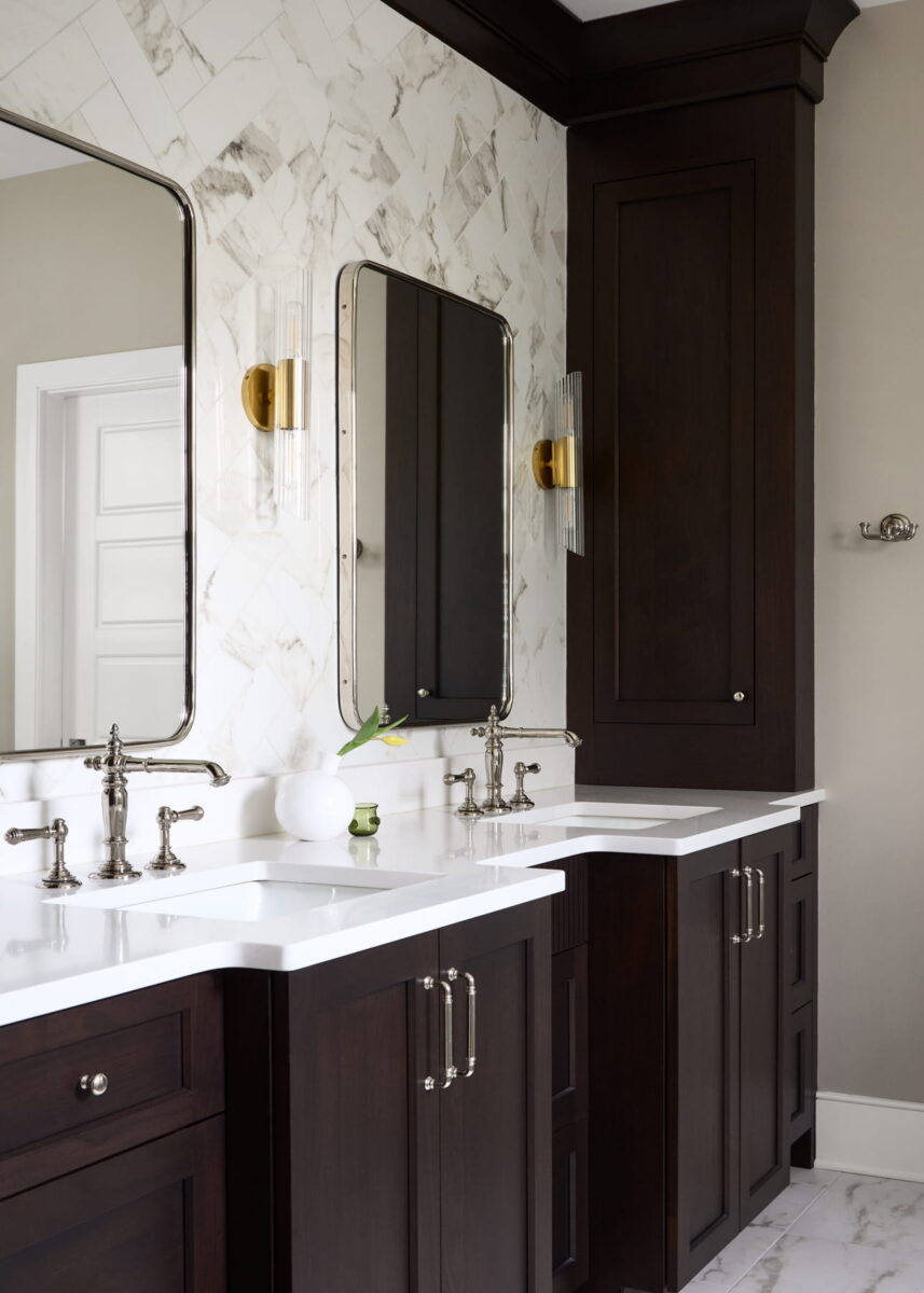 Modern bathroom with two rectangular mirrors, white marble wall, dark wood cabinets, double sinks with silver faucets, and gold wall sconces. White vase with a single flower sits on the countertop.