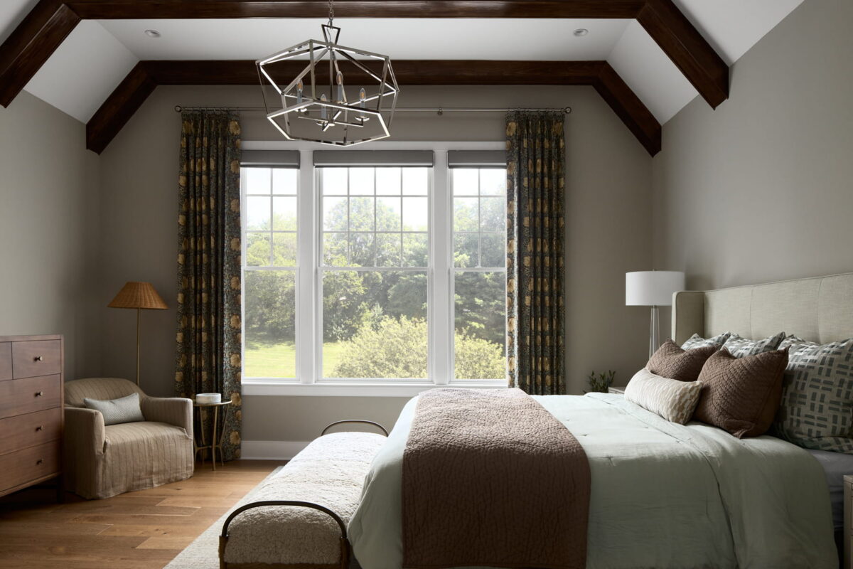A cozy bedroom with a large window, neutral walls, exposed wood beams, a bed with brown and blue bedding, an armchair, a dresser, and patterned curtains letting in natural light.