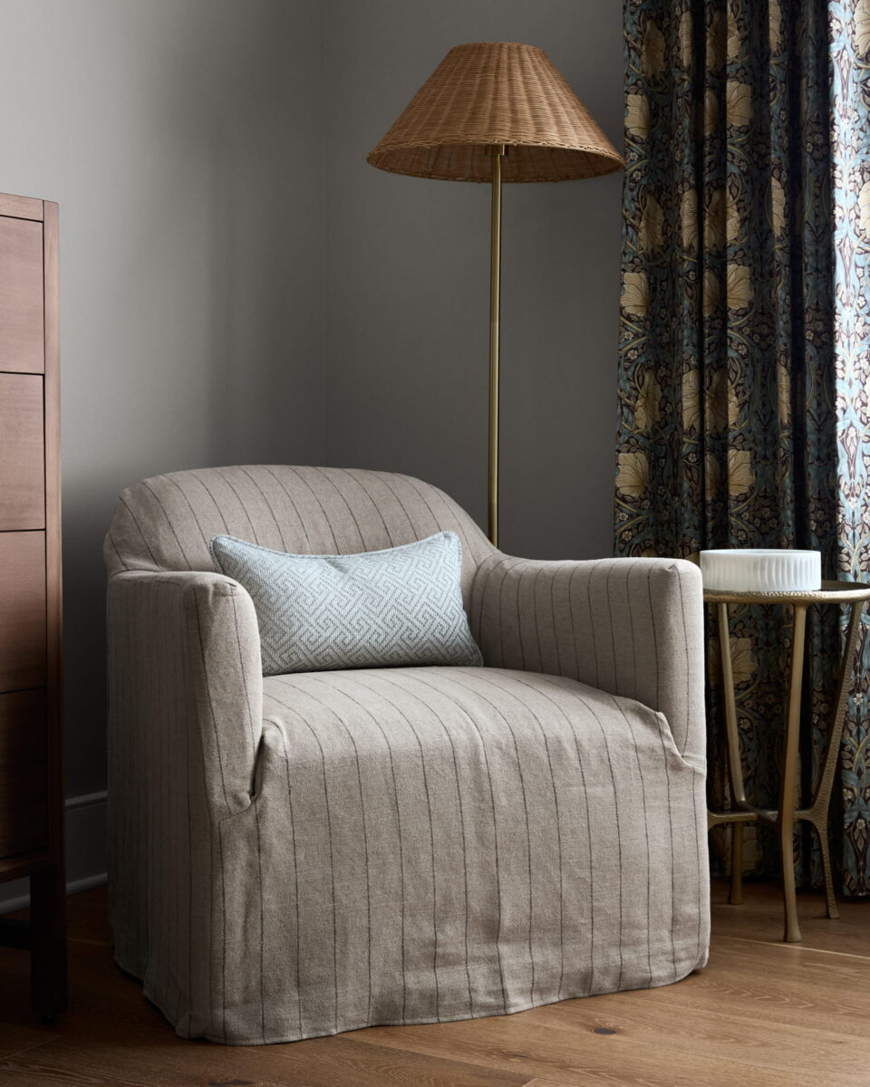 A beige armchair with subtle stripes and a blue patterned pillow sits next to a gold floor lamp, small round table, and floral curtains in a cozy, softly lit room.