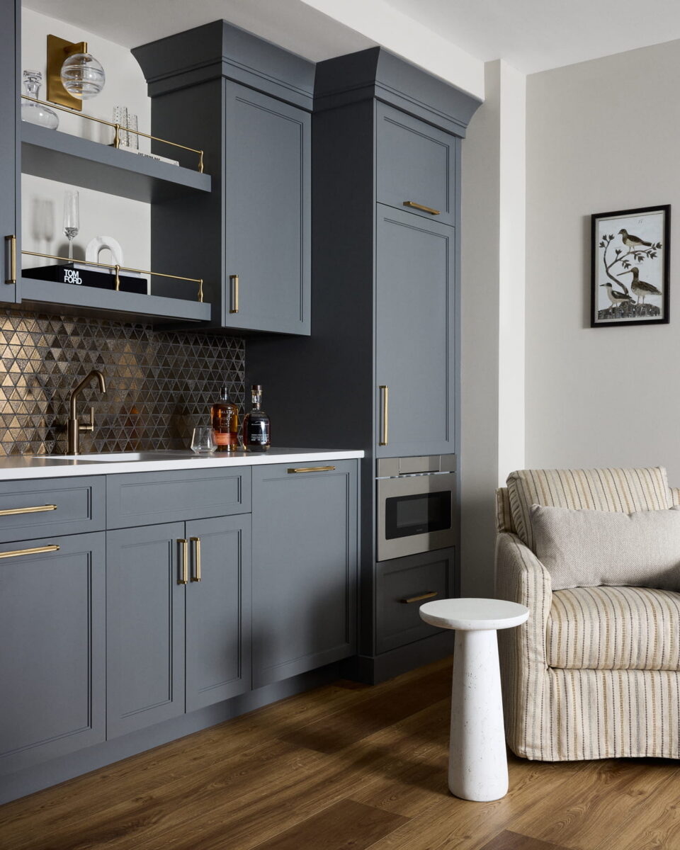 Modern kitchen with matte blue-gray cabinets, gold hardware, geometric tile backsplash, open shelves with glassware, and wood flooring. A beige striped armchair and small round white side table are nearby. Framed bird art hangs on the wall.