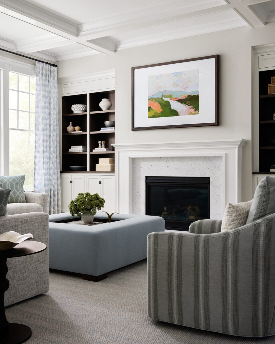 Modern living room with armchairs, ottoman, built-in shelves, fireplace, and abstract landscape art above mantle. Neutral tones with soft natural light from large window.
