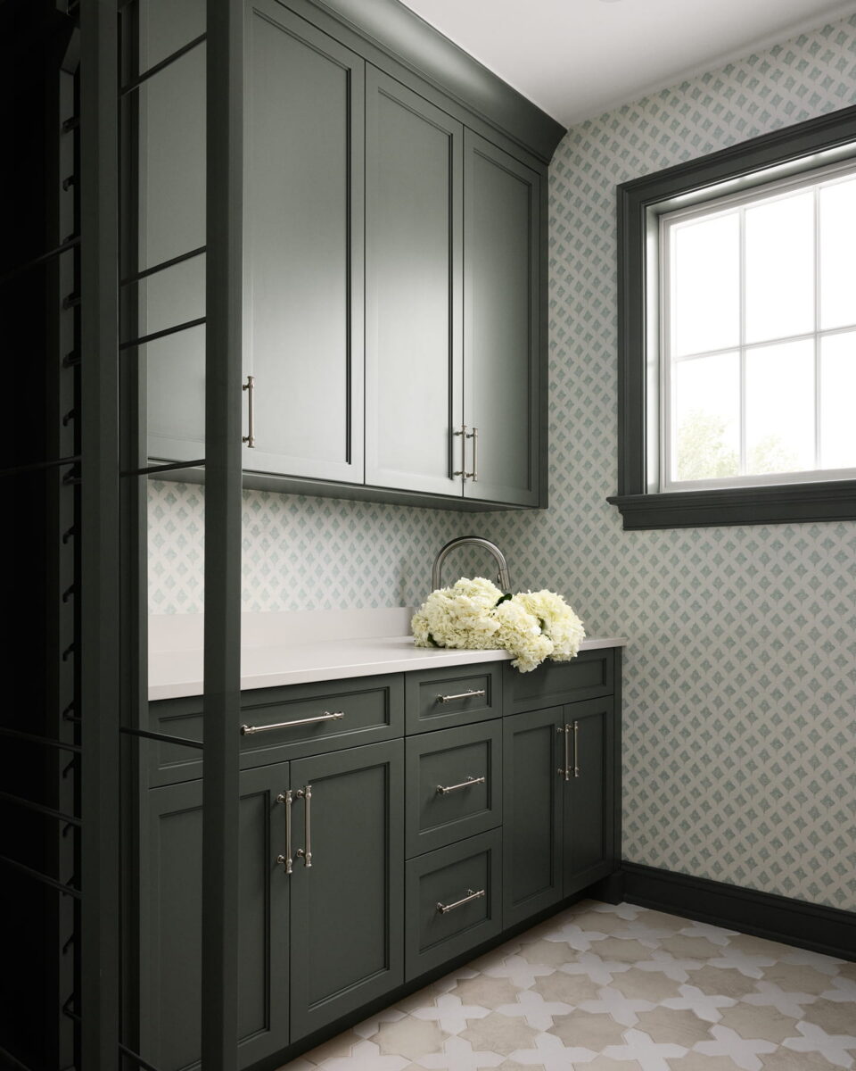 A laundry room with sage green cabinets, a white countertop, a bouquet of white flowers, patterned wallpaper, and a window with natural light.