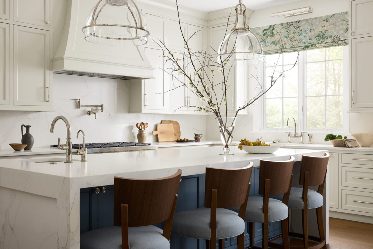 Bright, modern kitchen with white cabinets, a marble island with blue base, four wooden chairs, pendant lights, a branch in a vase, and a large window with floral curtains letting in natural light.