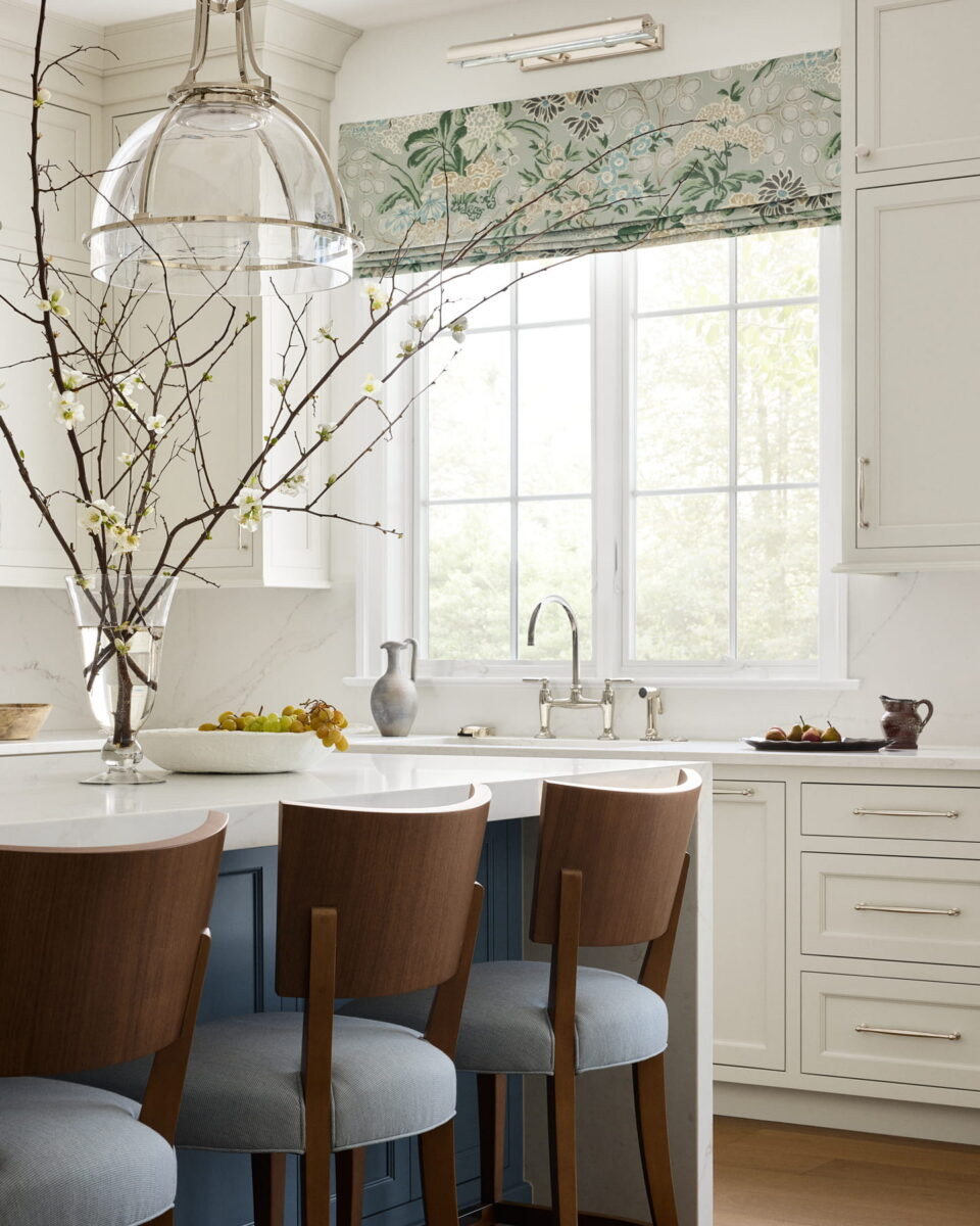 Bright kitchen with white cabinets, a window with a floral shade, a large vase with branches on the island, three wooden chairs with blue cushions, and fruit on the counter.