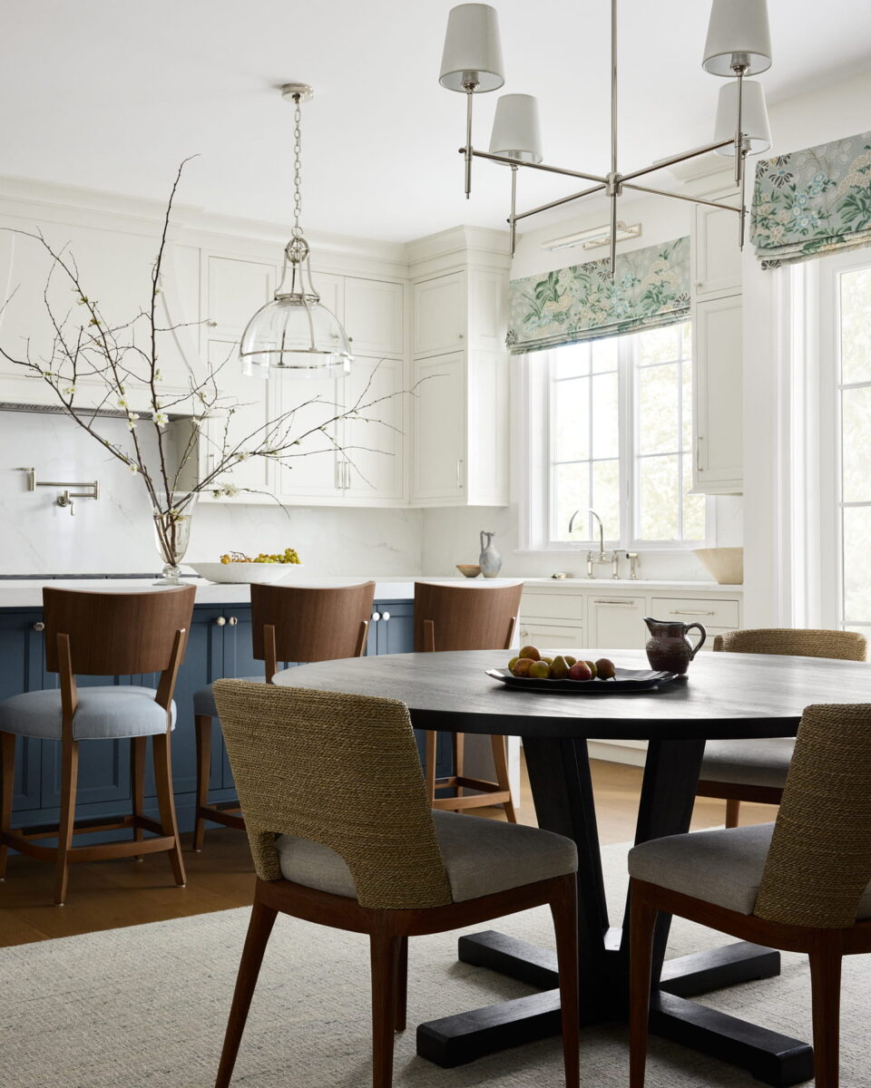 Bright kitchen with white cabinets, blue island, wooden chairs, a round dark table with beige chairs, large windows with floral shades, and a branch centerpiece in a vase on the island. Fruit and a teapot sit on the table.