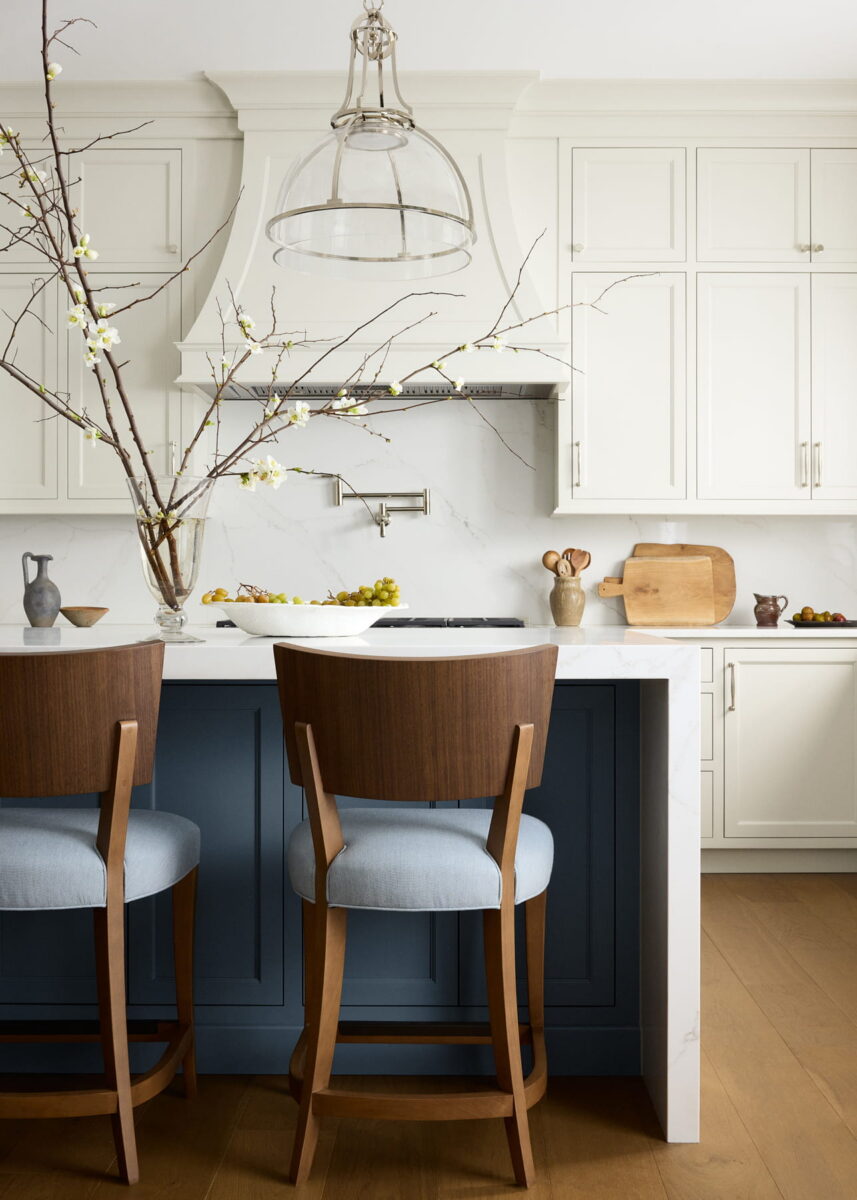 Modern kitchen with white cabinets, a marble backsplash, a blue island with two wooden chairs, glass pendant light, wooden cutting boards, and a tall vase with branches on the counter. Light wood flooring throughout.