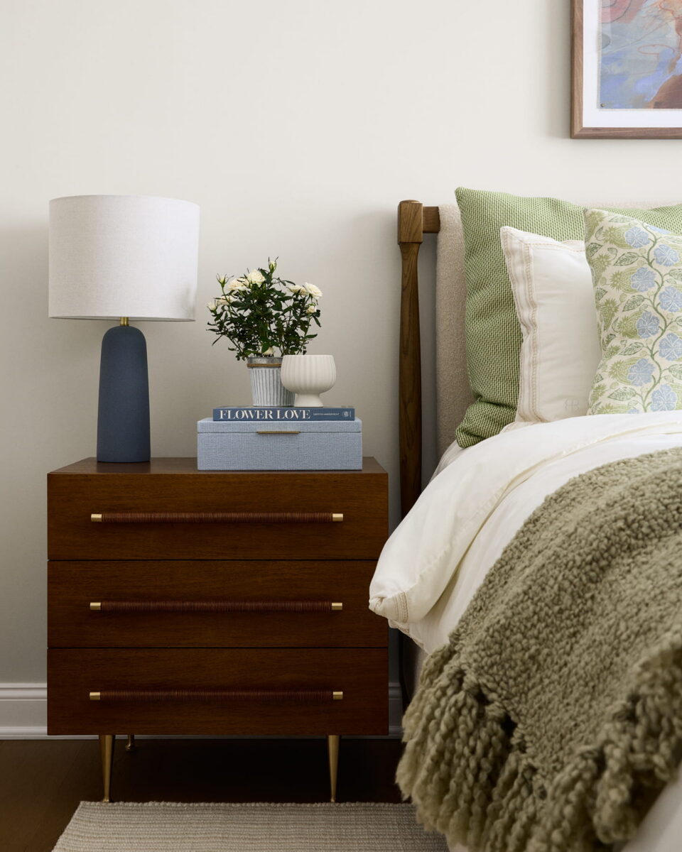 A wooden nightstand with three drawers holds a blue lamp, a vase of white flowers, and books. The bed nearby has green and white pillows and a textured green blanket. A framed picture hangs above.