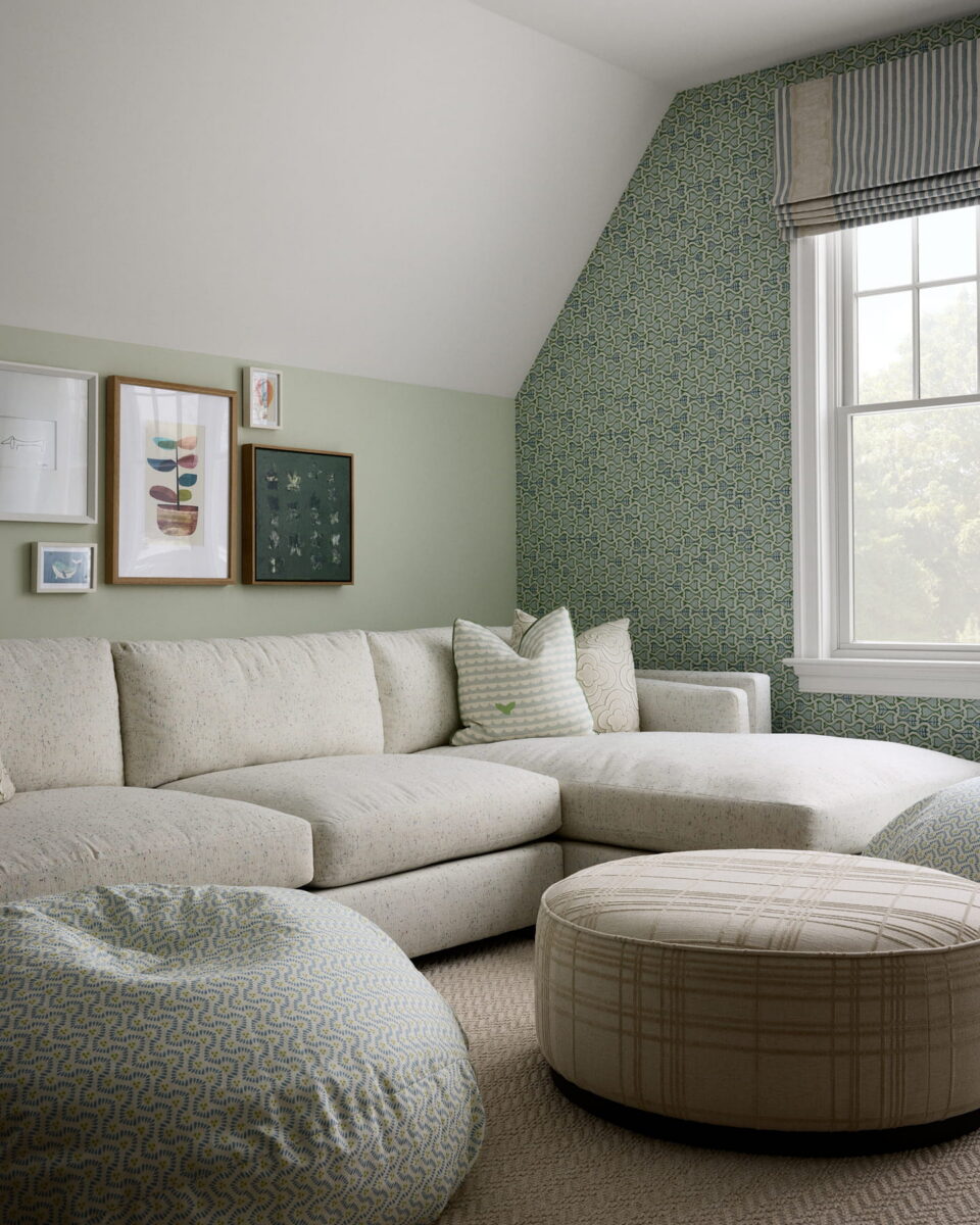 A cozy living room with a white sectional sofa, patterned bean bag, round plaid ottoman, green wallpaper, framed art, and a window with Roman shade letting in natural light.