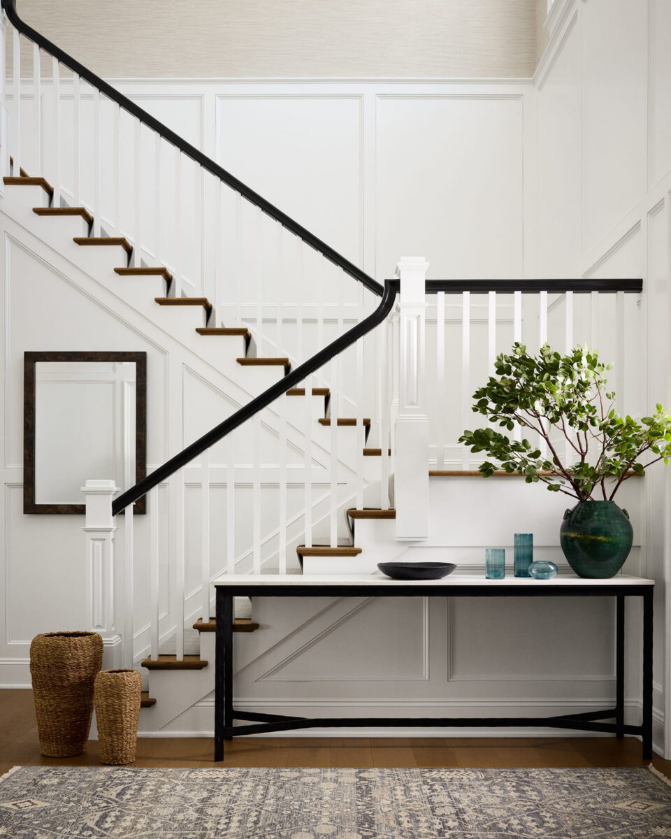 A modern, bright entryway with white paneled walls, a staircase with black railing, a black console table, a green plant in a vase, decorative bowls, glassware, a mirror, woven baskets, and a patterned rug.