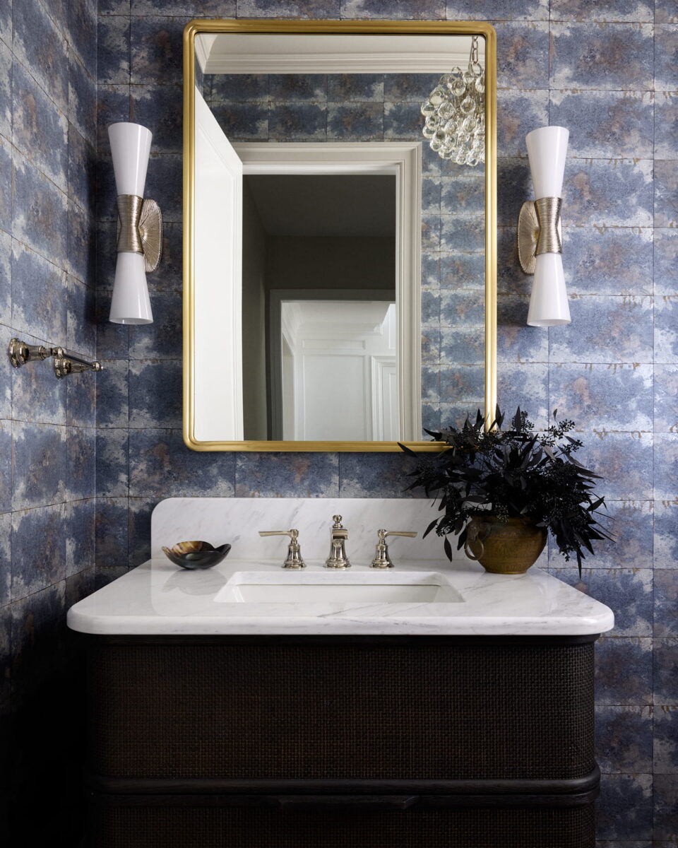 A bathroom vanity with a marble countertop, gold-framed mirror, modern sconces, and a vase of dark foliage against blue textured wallpaper. A door is reflected in the mirror.