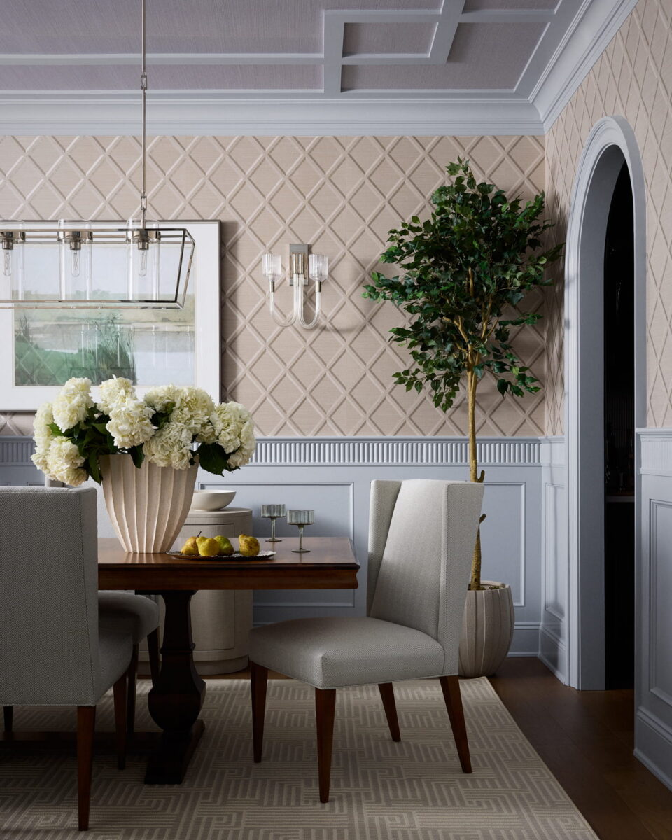 Elegant dining room with a wooden table, beige upholstered chairs, a large vase of white flowers, decorative wall panels, a potted tree, and a pendant light fixture. A bowl of pears sits on the table.
