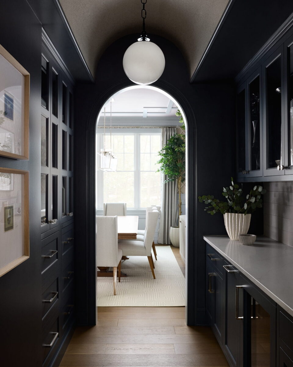 A dark, modern hallway with black cabinetry leads to a bright dining room with white chairs, large windows, and a potted tree. A round light fixture hangs from an arched doorway.