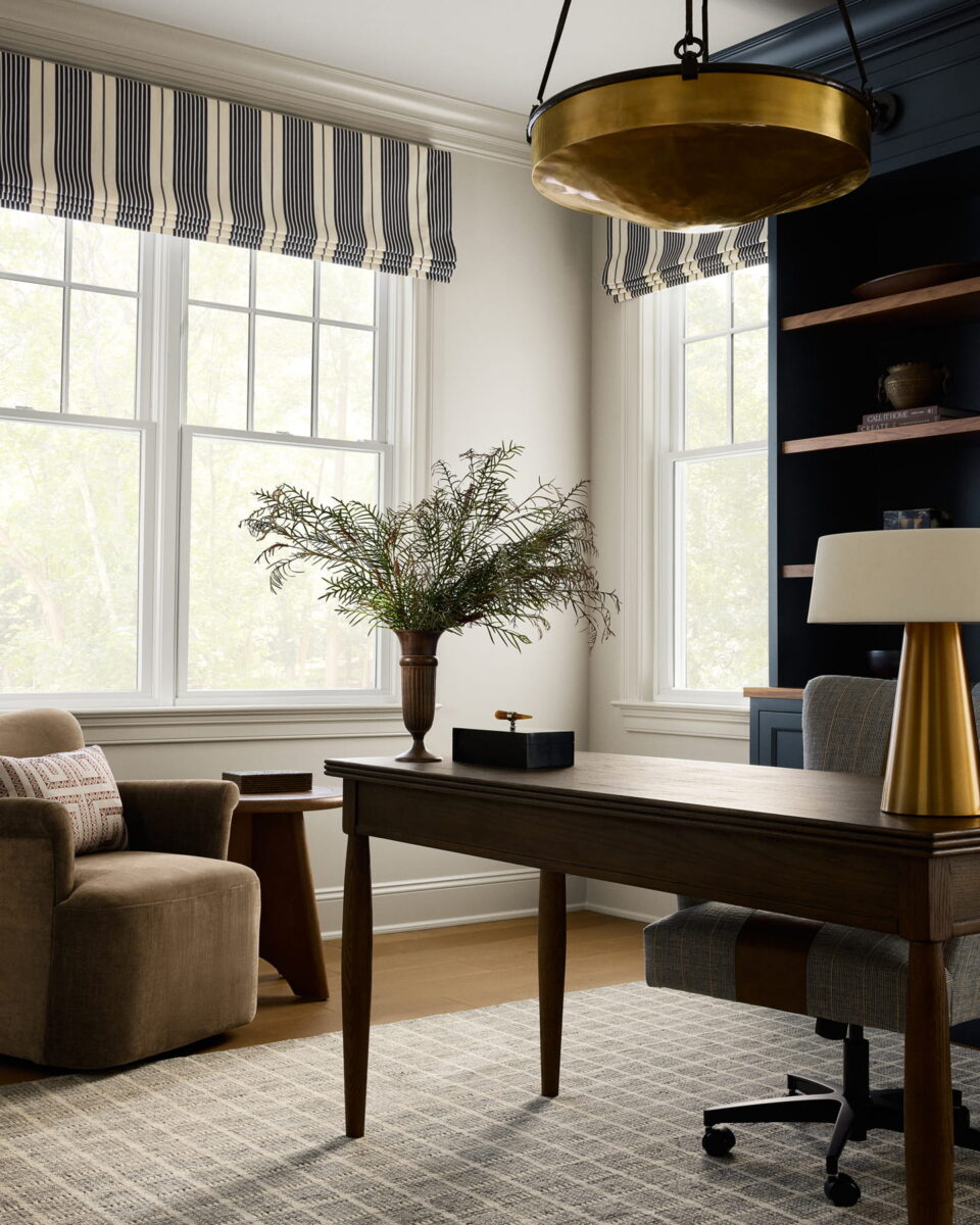 A stylish home office with a wooden desk, a modern lamp, a vase with greenery, a beige armchair, large windows with striped shades, and a gold chandelier overhead. Shelves with decor are visible on the right.