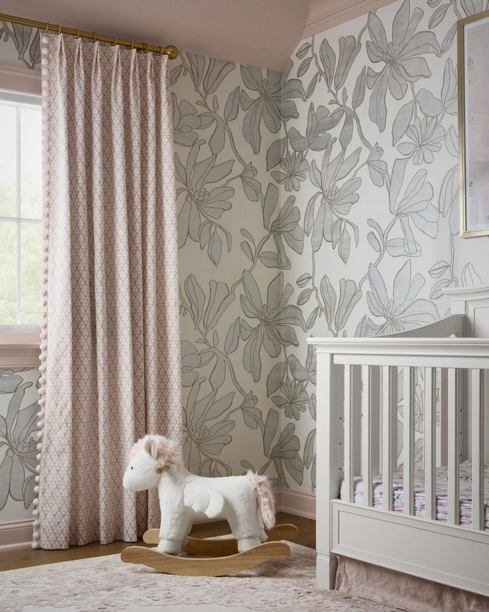 A nursery with floral wallpaper, a white crib, pink-patterned curtains, a pink rug, and a plush rocking horse shaped like a unicorn. Sunlight shines through the window, brightening the cozy, pastel-themed room.