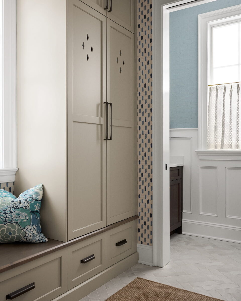 Built-in beige cabinets with black handles, a cushioned bench with patterned pillows, and geometric wallpaper. A doorway leads to a blue-walled bathroom with a vanity, white wainscoting, and a window with a pleated curtain.