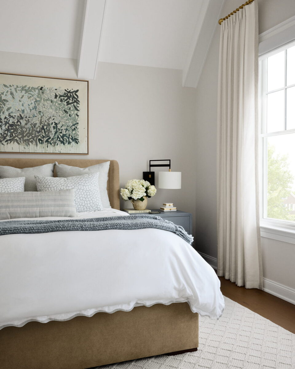 A cozy bedroom with a beige bed, white bedding, gray throw, textured pillows, nightstand with flowers, lamp, and books, light curtains, large window, soft rug, and a framed abstract artwork above the bed.