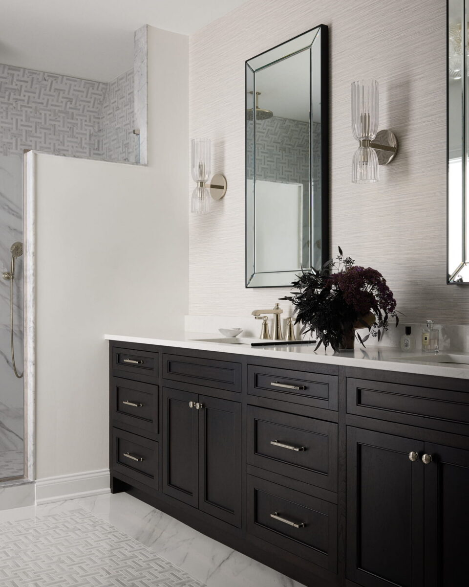 Modern bathroom with a dark wood vanity, white countertop, large wall mirror, glass sconces, and floral arrangement. Part of a walk-in shower with patterned tile is visible on the left.