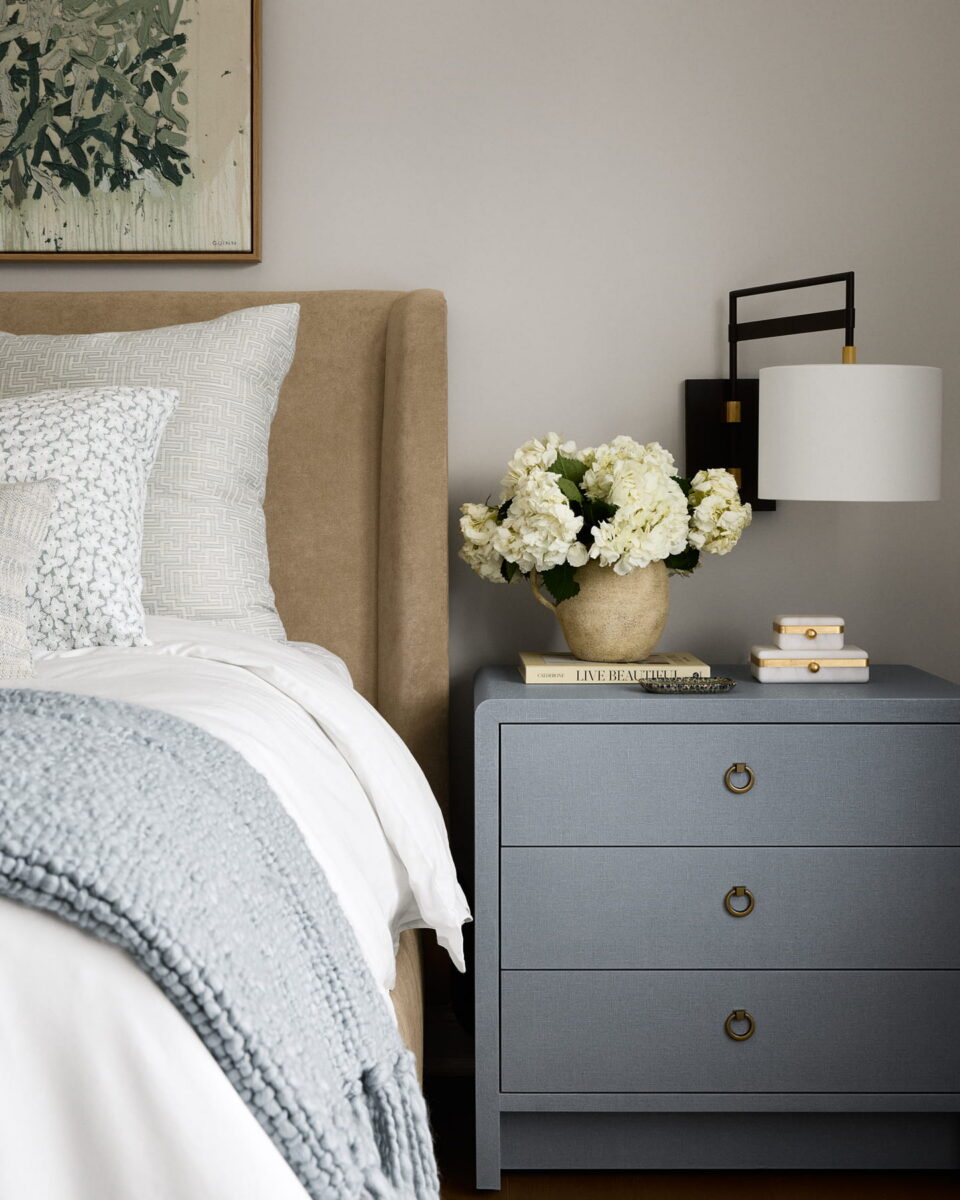A cozy bedroom scene with a beige upholstered bed, white bedding, a gray knit blanket, a blue nightstand holding stacked books, a bowl of white flowers, and a wall-mounted lamp above. Artwork hangs on the wall.