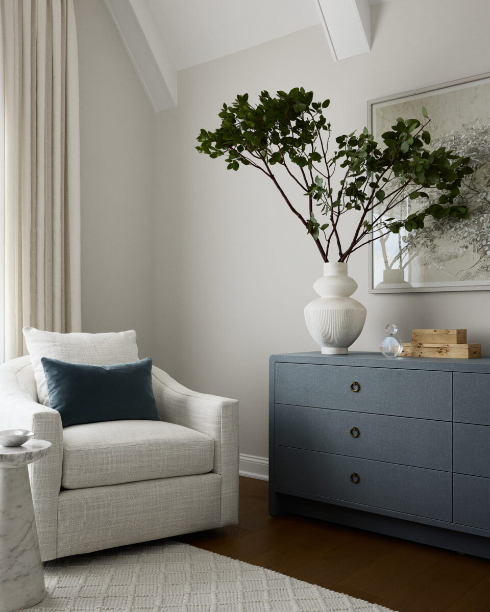 A cozy white armchair with a blue cushion sits beside a blue dresser topped with a white vase with green branches, a small sculpture, and decor. Neutral walls and soft curtains complete the modern, serene room.